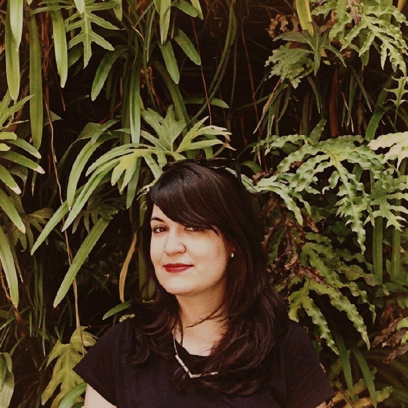 Photo d'une femme aux longs cheveux noirs et à un t-shirt noir avec des plantes en arrière-plan